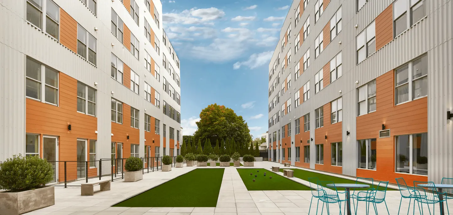 A modern courtyard view between orange-accented buildings under a clear blue sky, featuring green grass and trees.