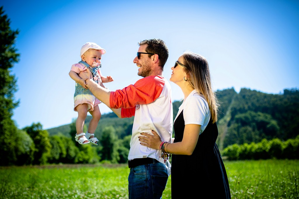 A joyful family enjoys a sunny day in a lush green field, with the father lifting their baby into the air while the mother watches and smiles. The bright blue sky and natural backdrop highlight the happiness and togetherness of the family, celebrating precious moments in nature.