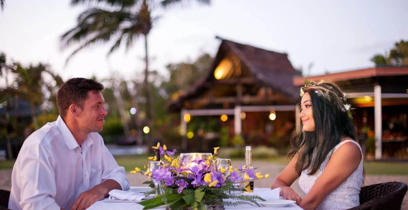 Couple enjoying a romantic beachfront dinner at Uprising Beach Resort in Pacific Harbour, Fiji. Lit resort building behind them.