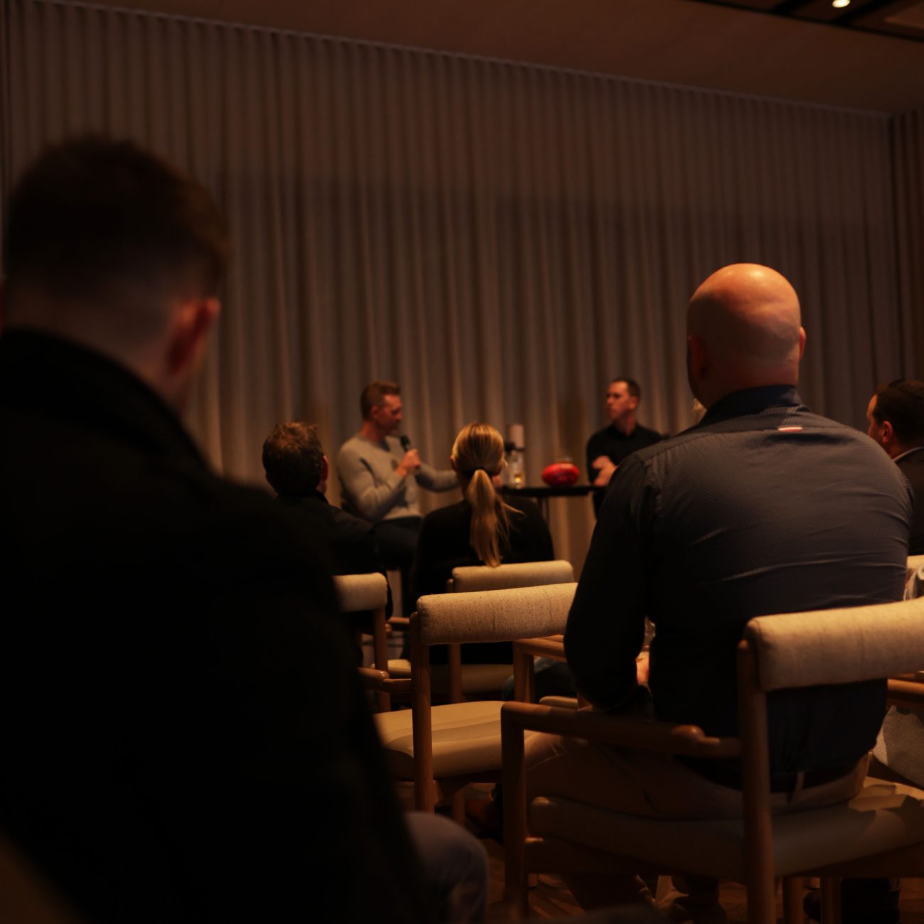 Audience watching a panel discussion between two men seated on stools by a curtained window