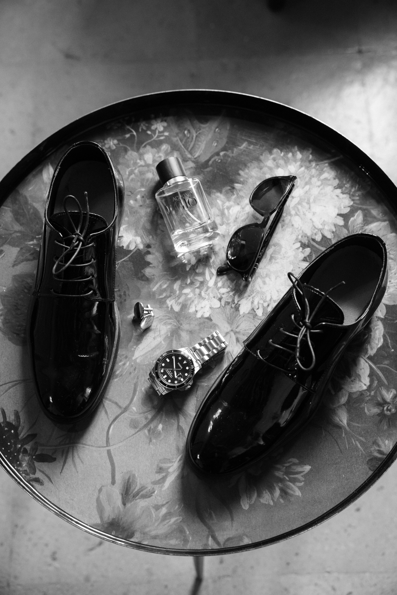 Dettagli matrimoniali dello sposo: scarpe nere, profumo, occhiali da sole e orologio su tavolo con texture bianca e nera