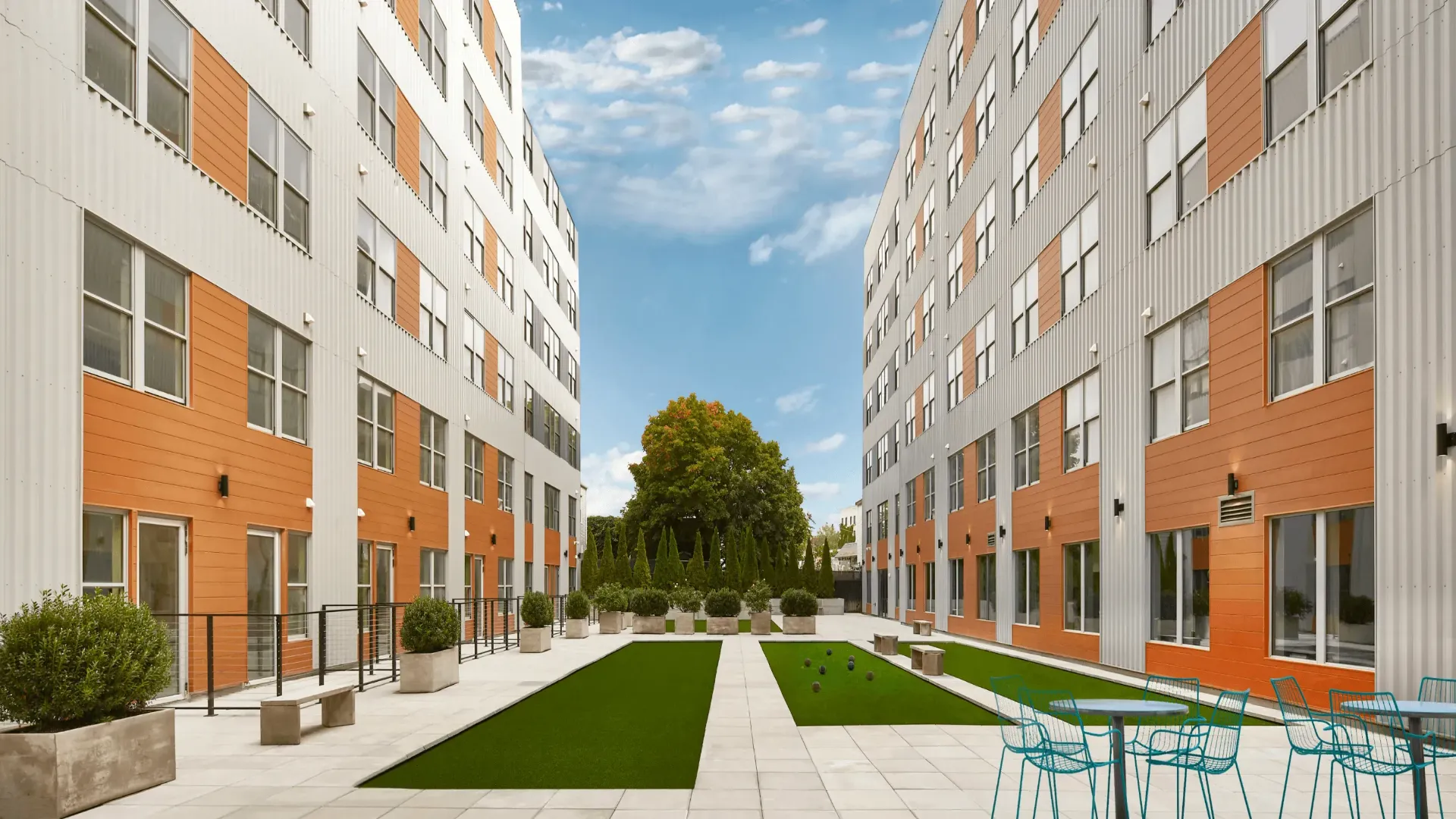 A modern courtyard view between orange-accented buildings under a clear blue sky, featuring green grass and trees.