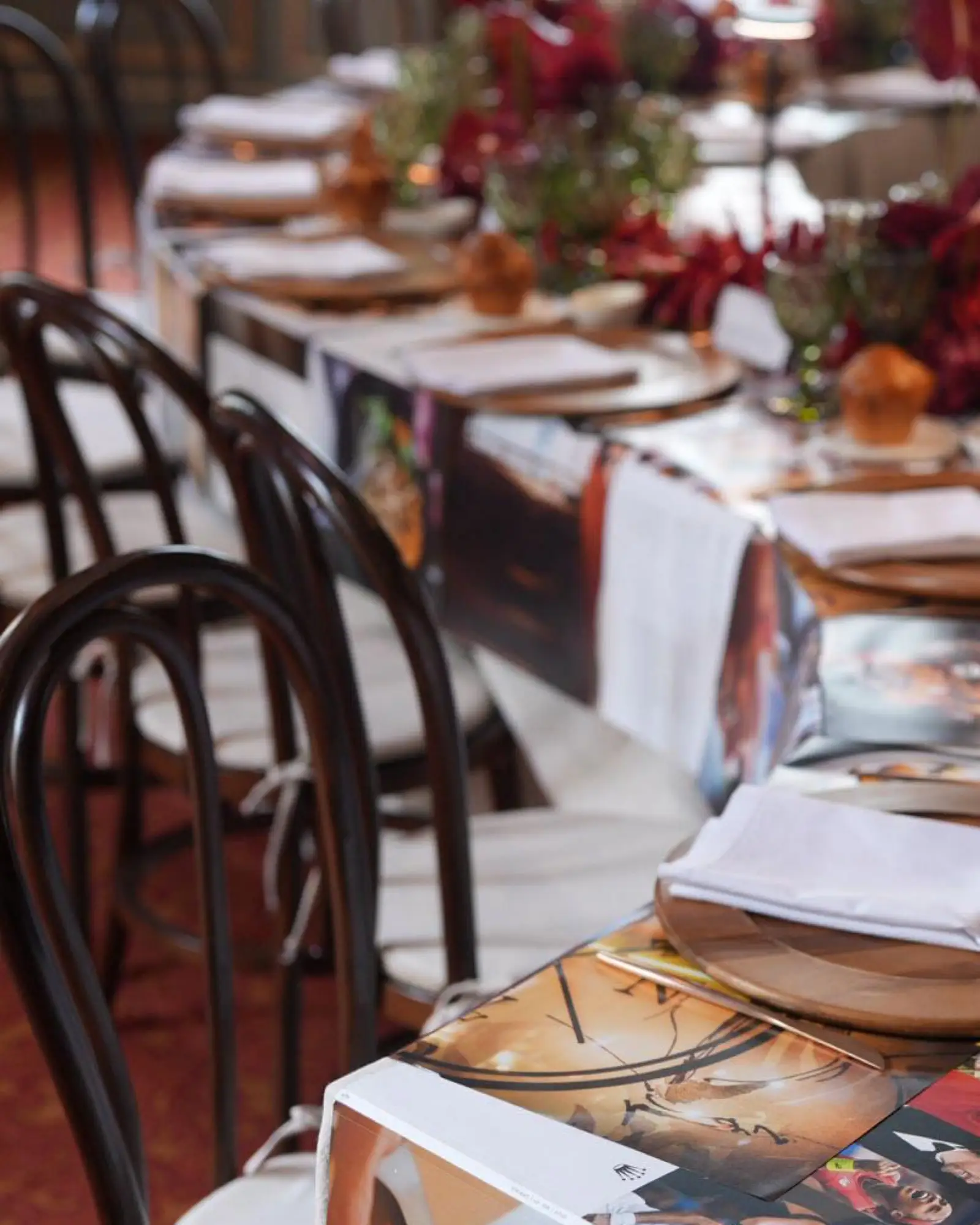 Sillas Thonet de madera oscura junto a mesa de banquete con mantel impreso, bajoplatos de madera y centros rojos.
