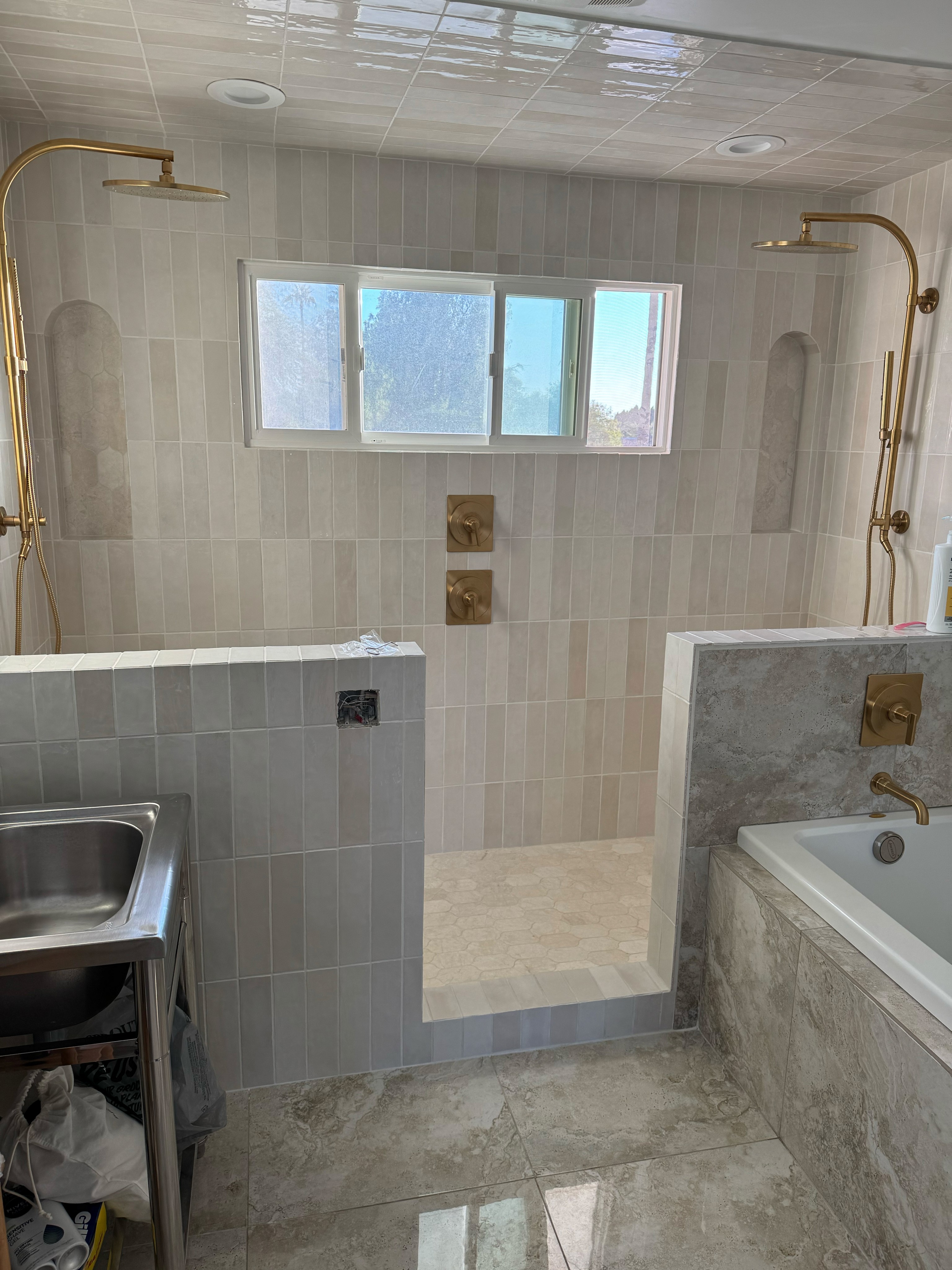 Custom walk-in shower with vertical beige subway tile walls, dual brass shower fixtures, and built-in niches, next to a tile-wrapped bathtub.