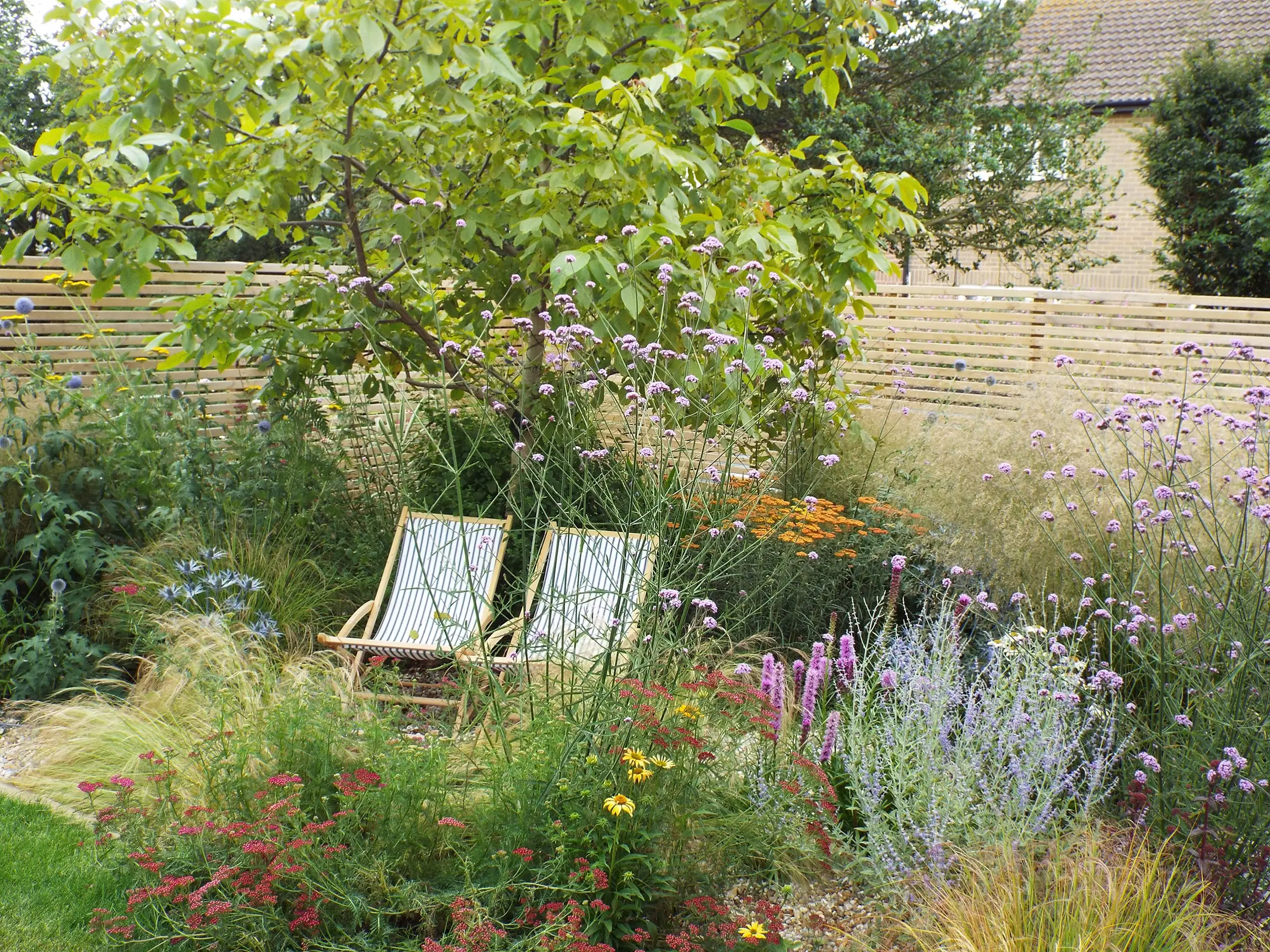 A serene garden scene featuring two deck chairs under a tree, surrounded by colorful flowers and greenery.