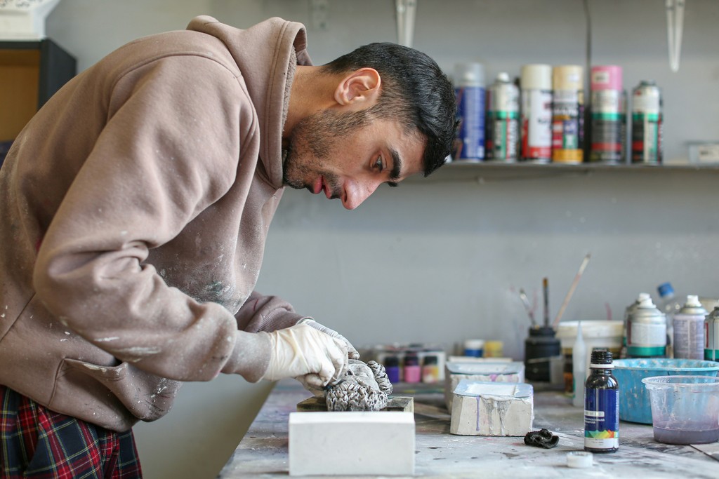 An artist intensely works on a sculpture in a cluttered studio, surrounded by various art supplies including spray paint cans, brushes, and bottles. The individual, dressed in a hoodie and gloves, is deeply focused on crafting a detailed piece, capturing the dedication and creativity involved in the artistic process.