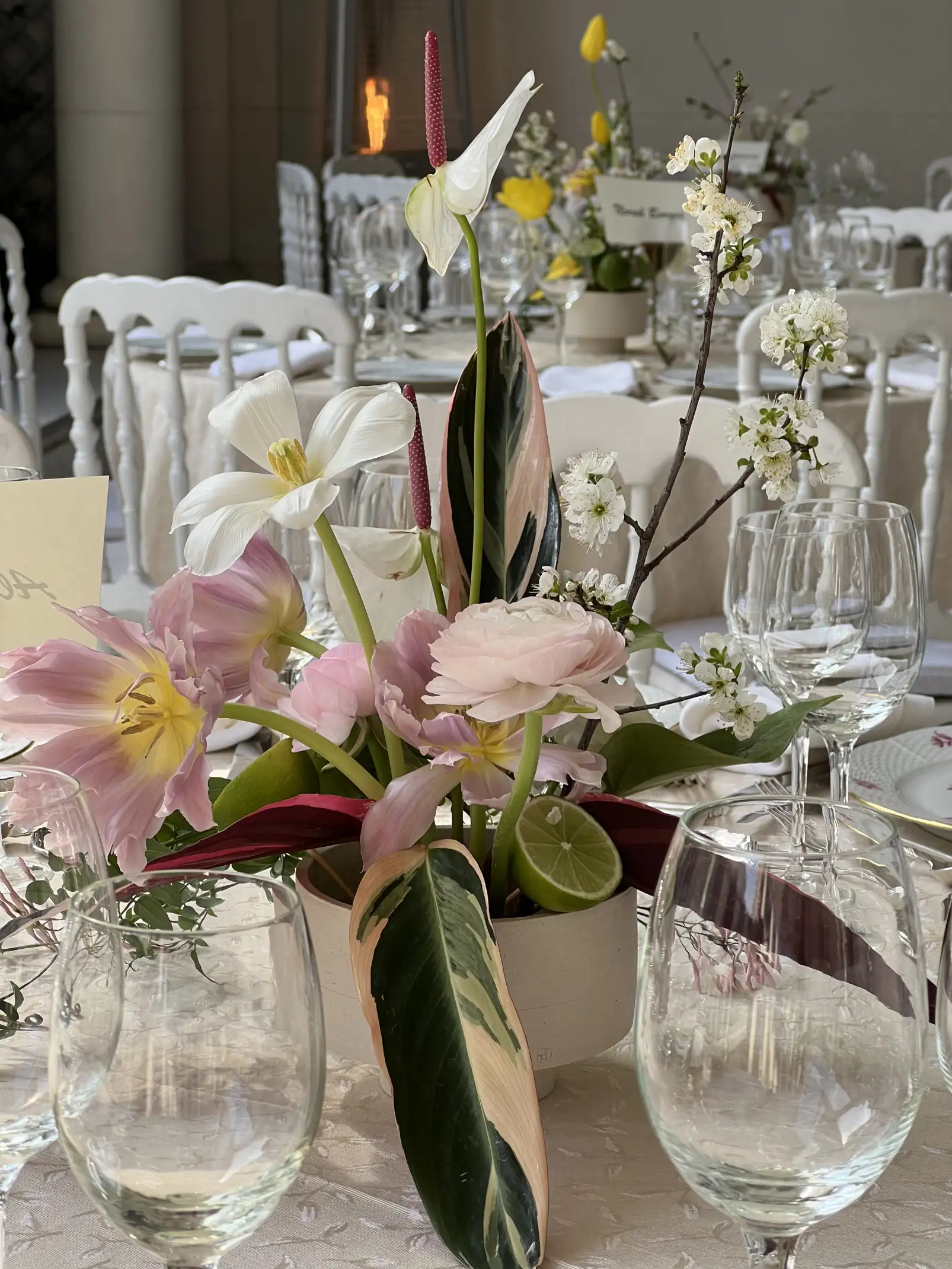 Centro de mesa primaveral con anturios blancos, tulipanes rosados, ranúnculos, flores de cerezo y hojas variegadas, dispuesto en un cuenco de cerámica sobre una mesa vestida en tonos crema con copas de cristal y vajilla blanca.