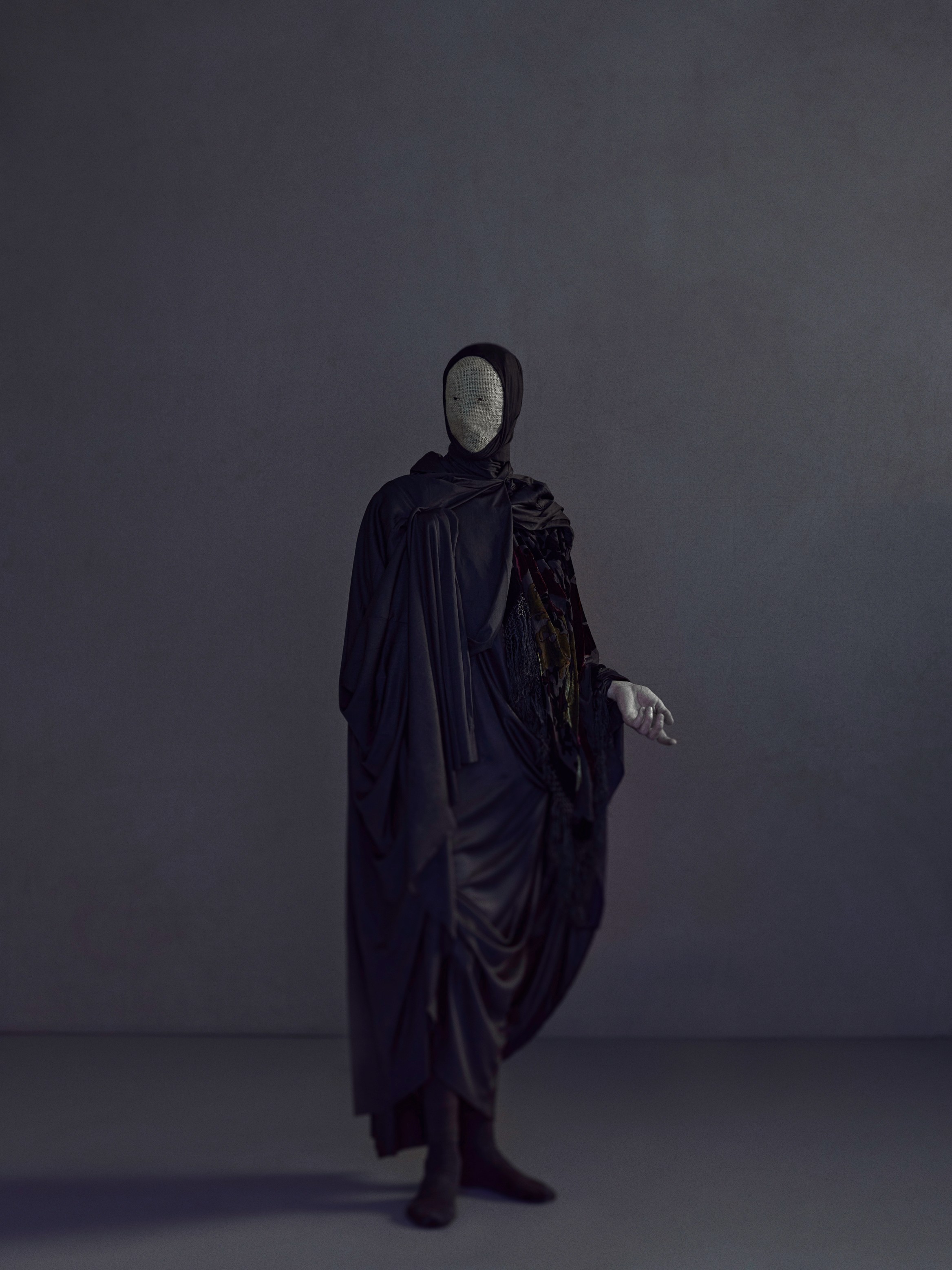 An ominous ancient masked figure stands within a minimal textured space. 