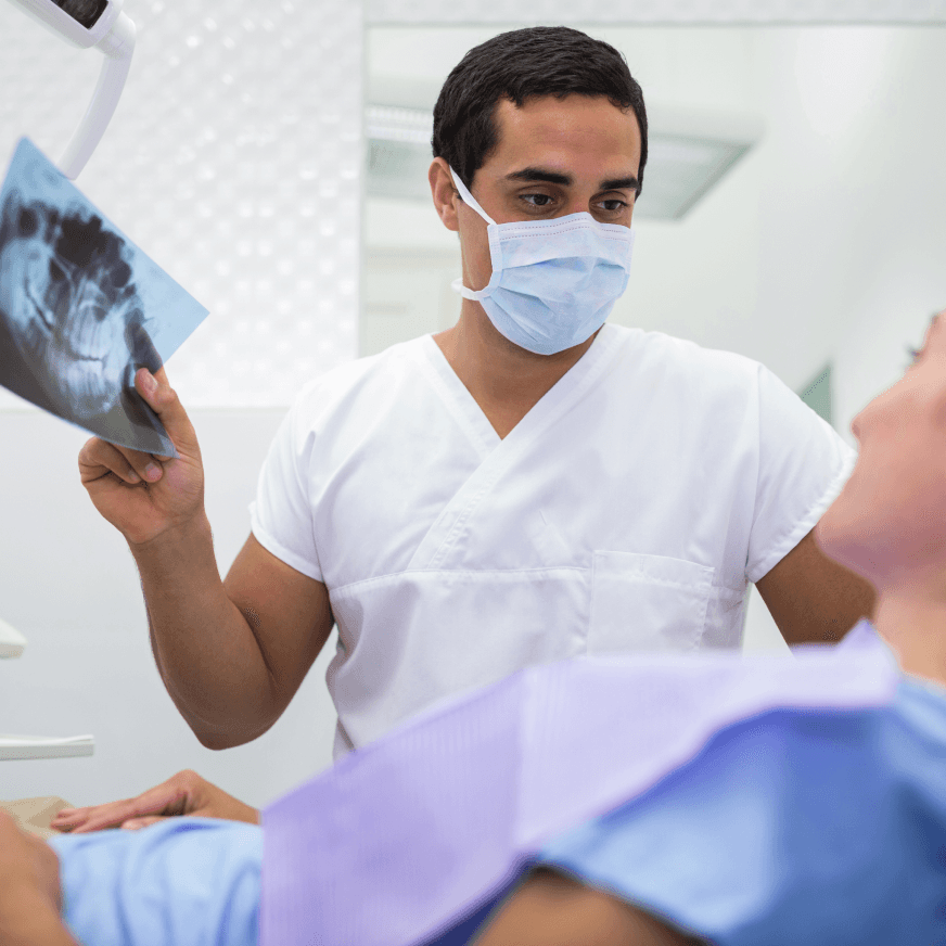 A dentist wearing a mask showing a dental X-ray to a patient in a dental chair, discussing the findings.