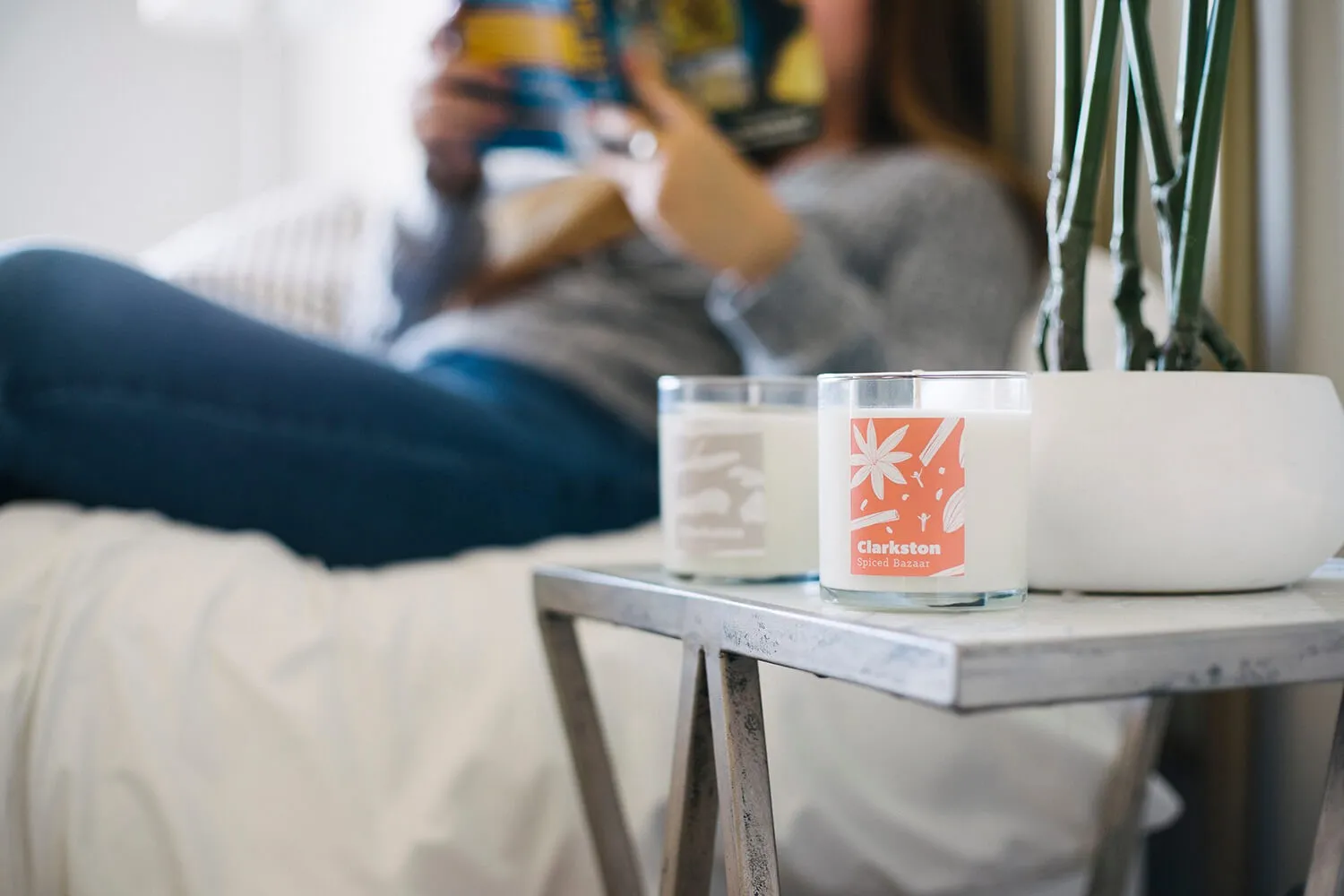 Set of candles on a side table in a bed room setting with a person reading in the background on bed.  