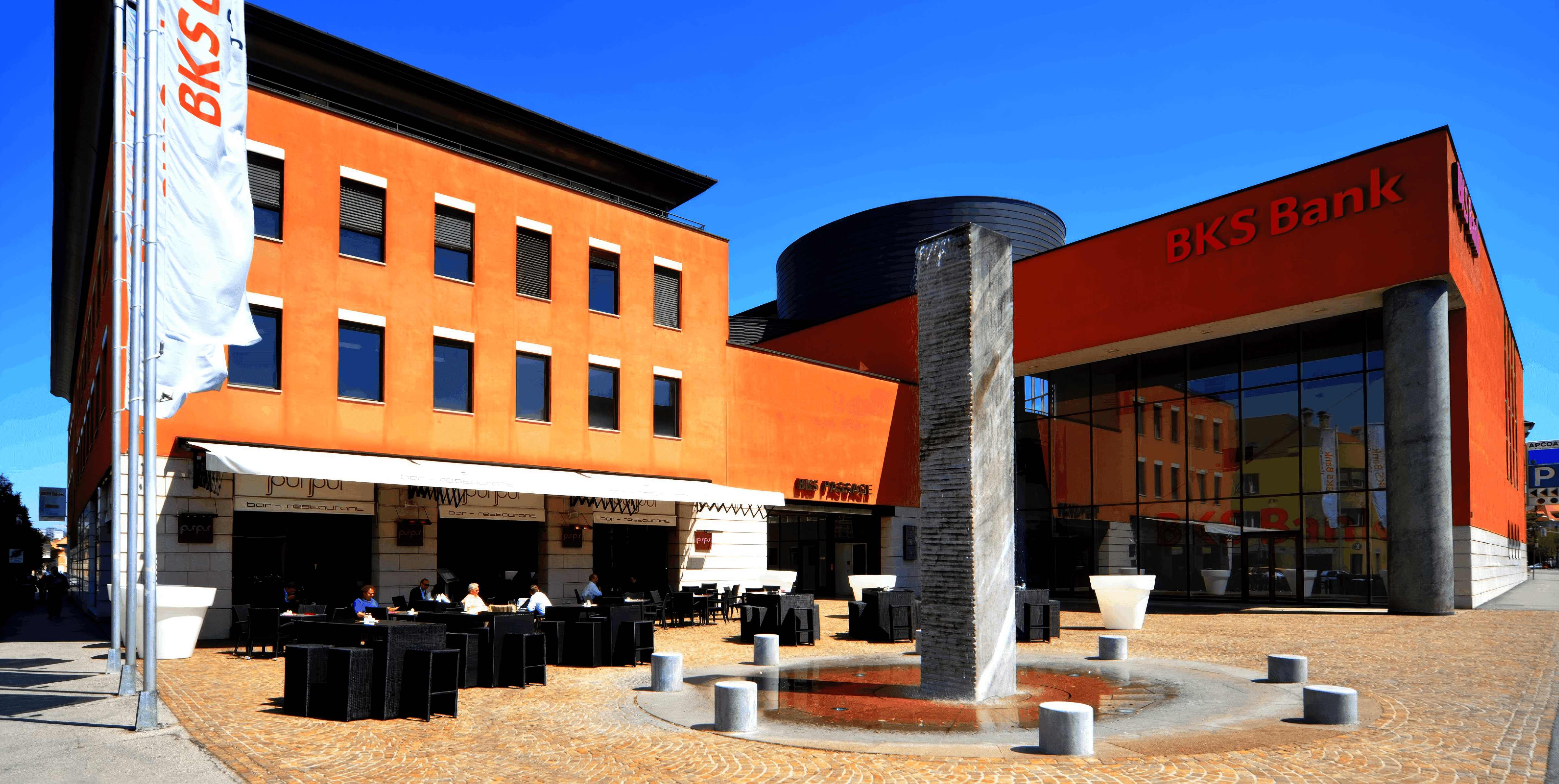 MW Interior - BKS Bank Hauptsitz mit markanter orangeroter Fassade, Glasfront und belebtem Vorplatz mit Brunnen in der Klagenfurter Innenstadt" Beschreibung: Markanter BKS Bank-Standort mit charakteristischer Fassade und belebtem Stadtplatz – ein anspruchsvoller Referenzkunde, für dessen Inneneinrichtung maßgeschneiderte Objektlösungen realisiert wurden.