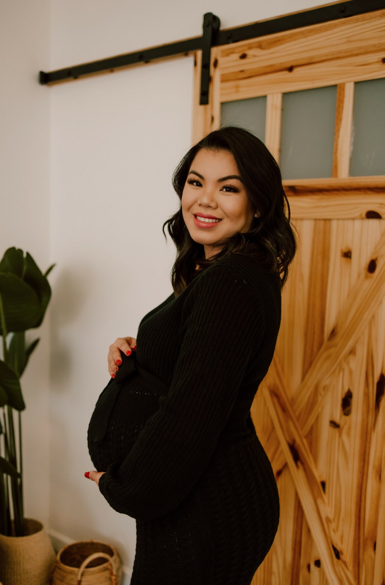 A pregnant woman, dressed in a black sweater, stands gracefully in front of a wooden door.