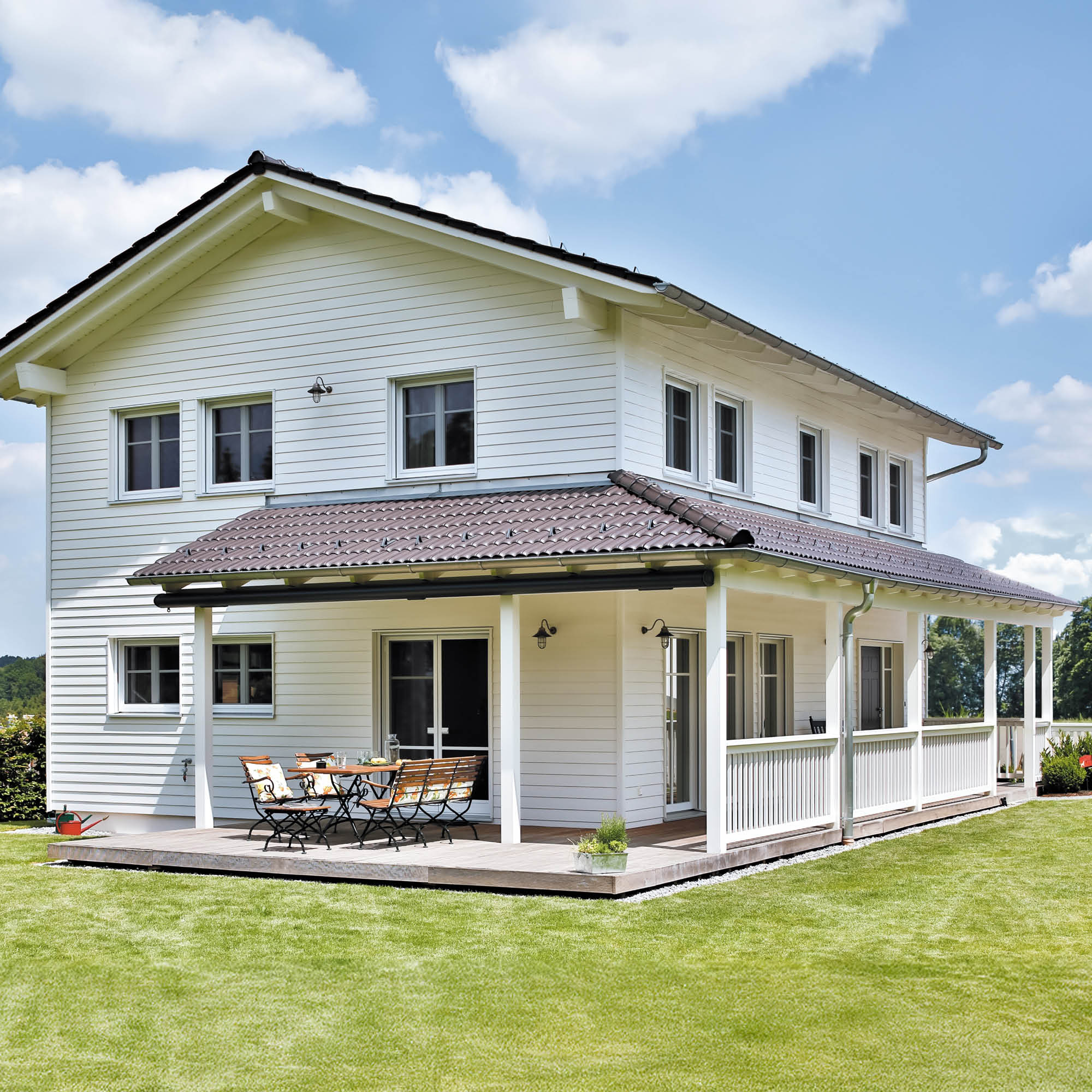 Ein weißes Doppelhaus mit einer umlaufenden Veranda und einem hölzernen Terrassenbereich mit Sitzmöbeln an einem sonnigen Tag mit blauen Wolken.