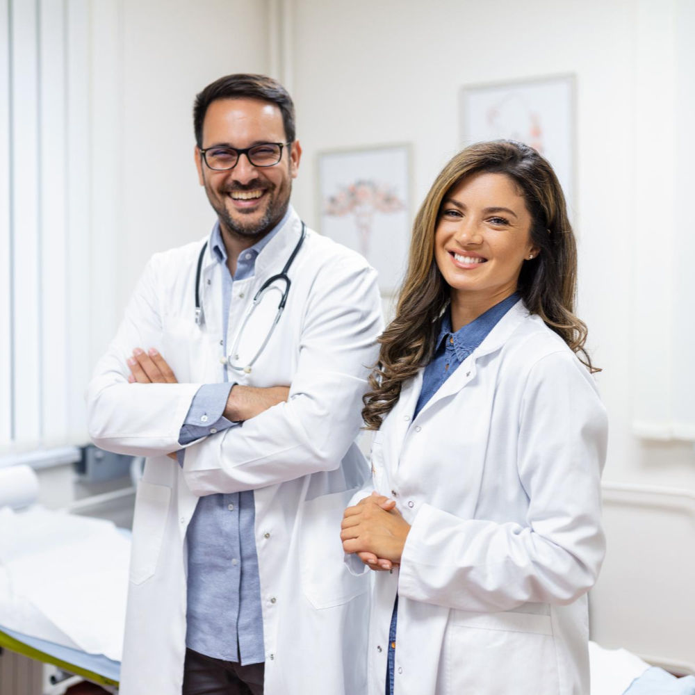 Two doctors in white lab coats and stethoscopes standing confidently in a clinical setting with medical posters and a bed in the background