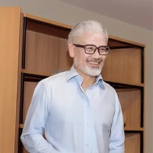 A person with short white hair and a beard, wearing glasses and a light blue shirt, is standing in front of a wooden bookshelf in a brightly lit room.