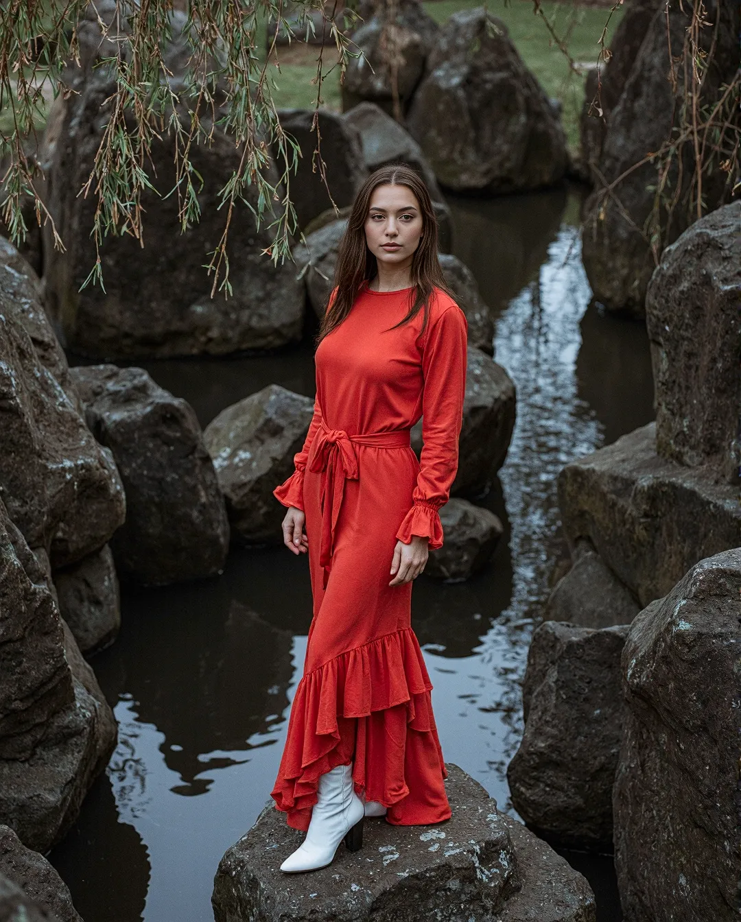A vibrant red dress with ruffled details stands out dramatically against dark stone formations and still water, creating a moody editorial scene.