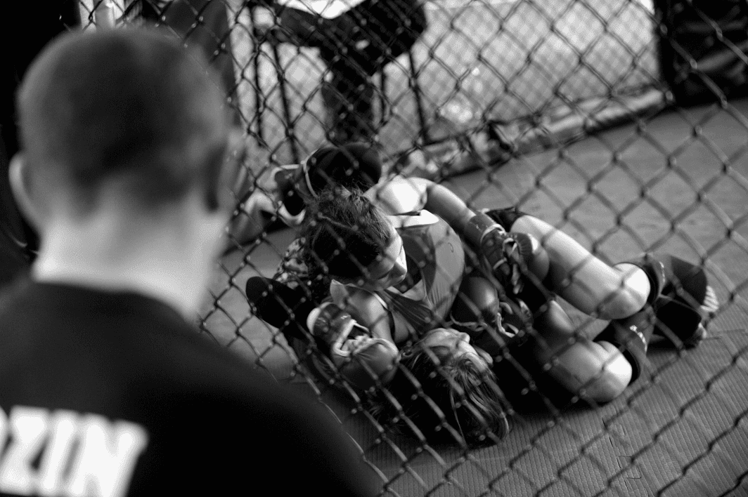 Two female MMA fighters grappling on the mat inside a cage while a referee watches through the fence.