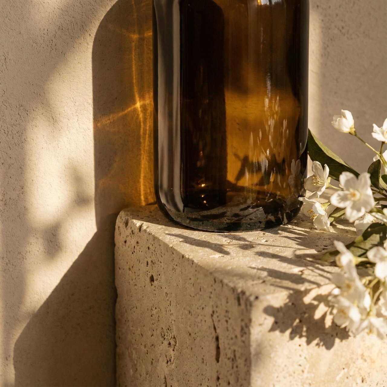 Brown glass bottle reflecting sunlight on a textured stone surface, surrounded by delicate white flowers. The scene feels warm and tranquil.