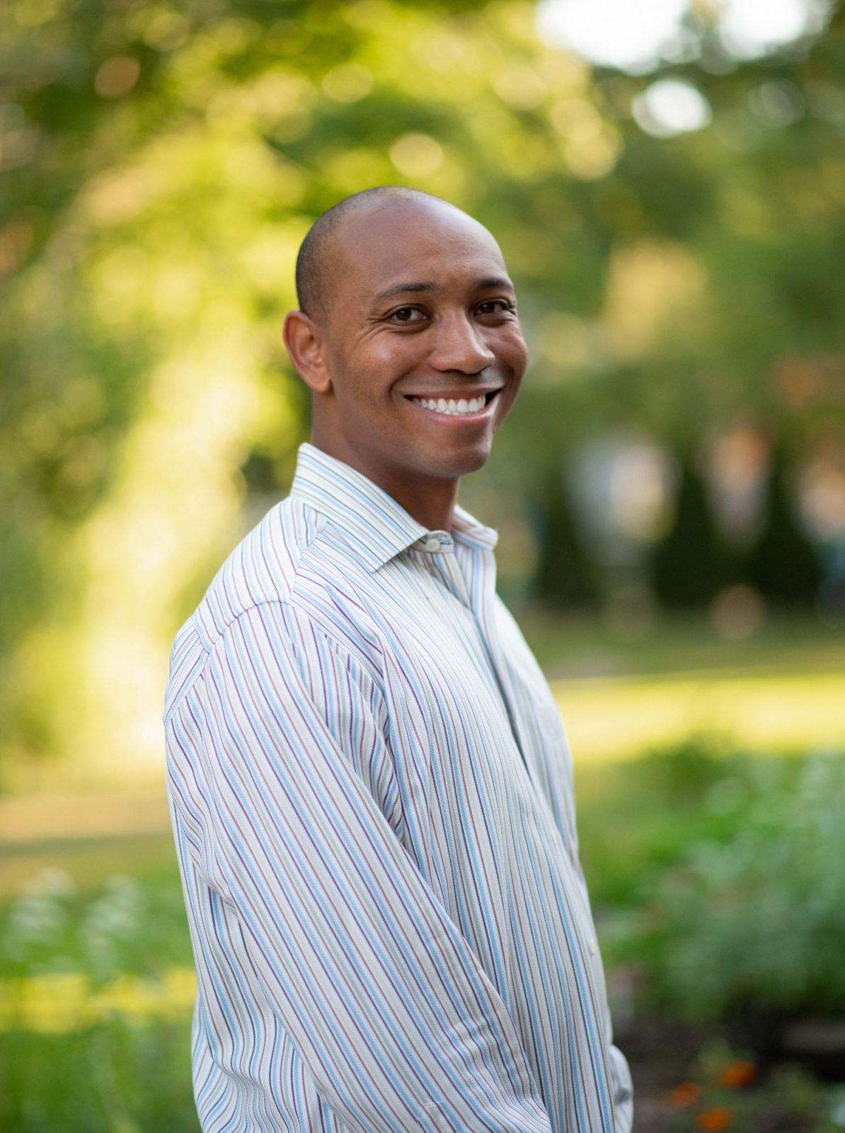 A man smiles while standing outdoors in a park, surrounded by greenery and sunlight.