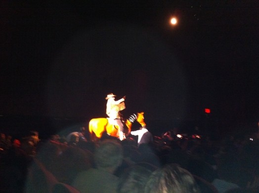 A photo of a man sitting on an artificial brown horse with his head attached by a string to the body. A group of people is watching the man while he is lit by a theatre lamp.