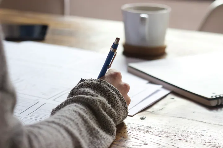 woman's hand writing with a pen to paper in the morning light, a coffee cup and notebook in the background