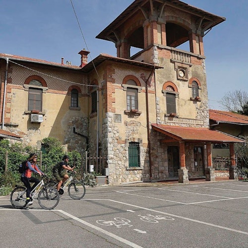 Two people ride bicycles past a rustic, multi-story building with arched windows and a red-tiled roof.