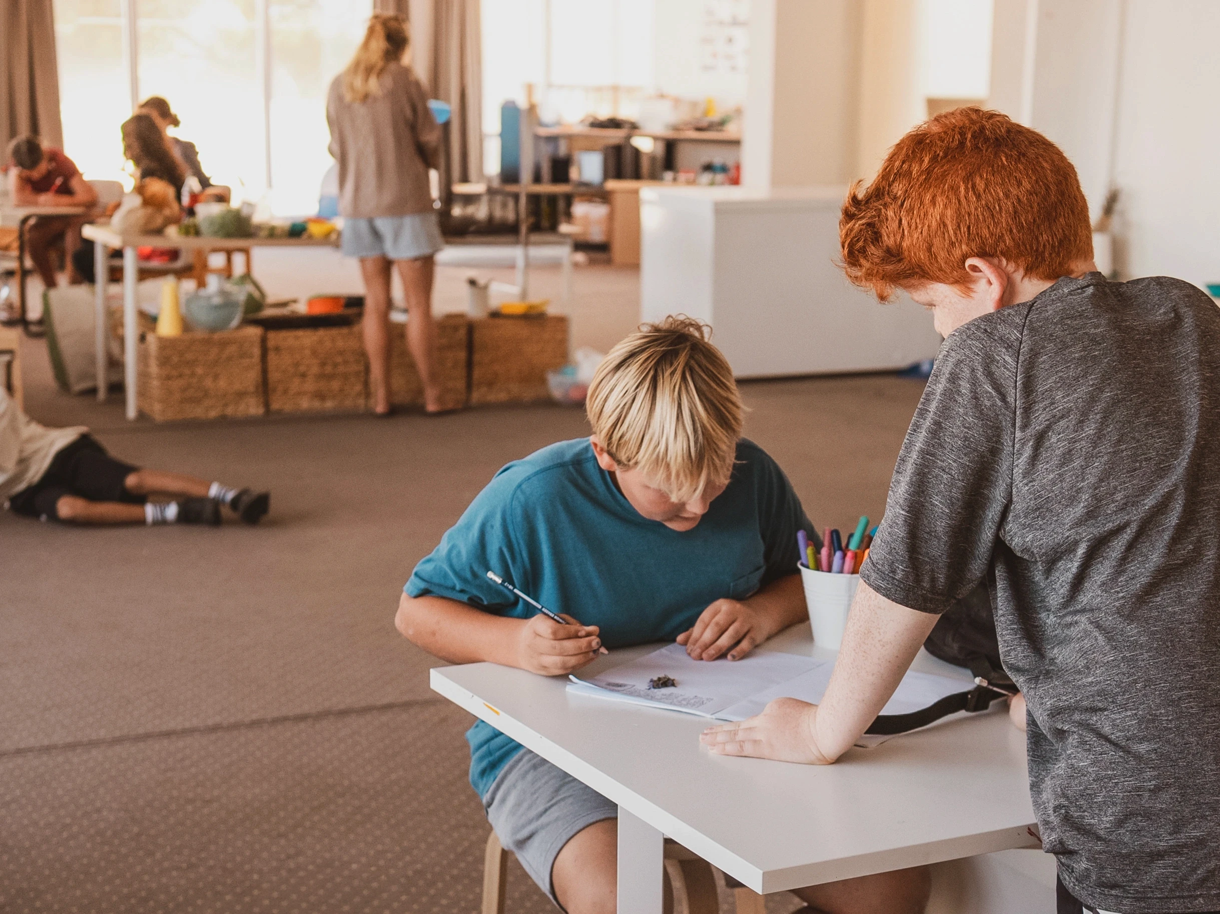 Two children at a white table in a classroom, one working in a notebook while the other looks on; art supplies and other people in the background.