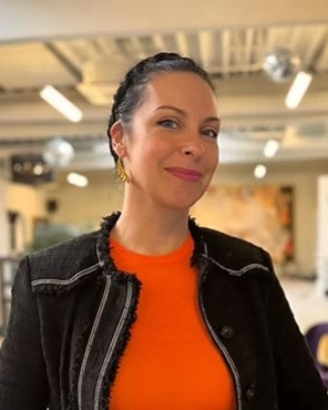 Eine junge Frau, mit einem orangenen T-Shirt und schwarzer Jacke, frontal in die Kamera lächelnd.