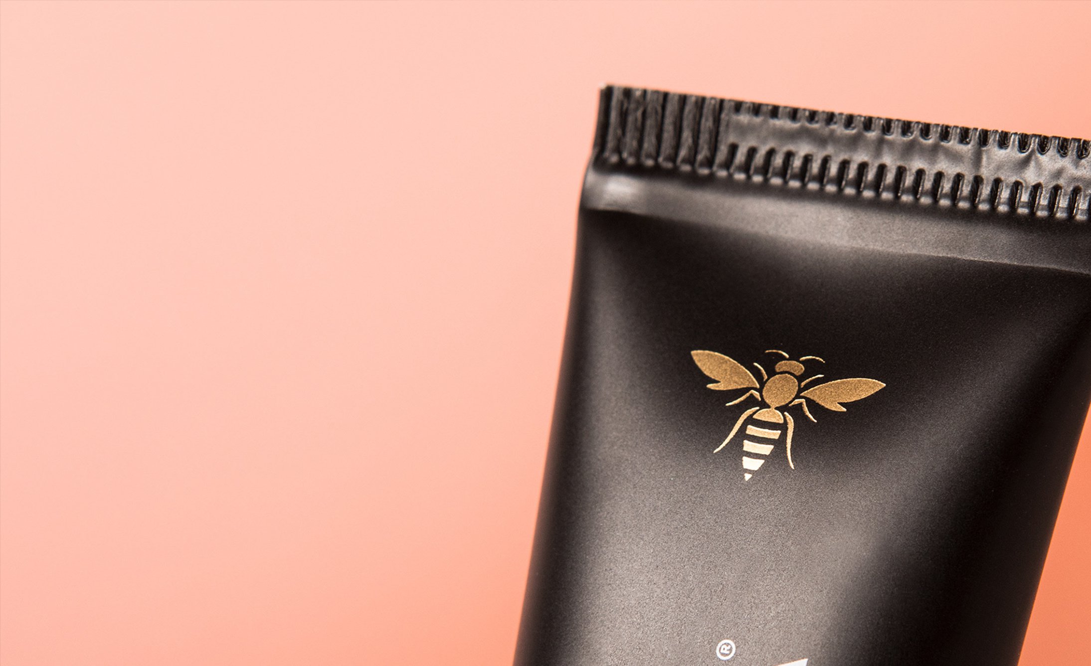 Black tube packaging with a gold bee logo on a pink background.