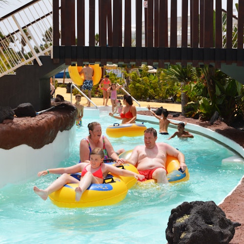 成年人和儿童在水上乐园的懒人河上乘坐充气管漂流,背景中有郁郁葱葱的绿地和其他人。