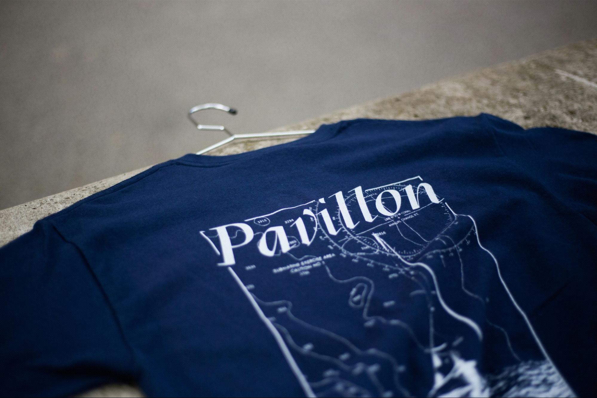 A navy blue t-shirt with a white design on its back. The design features a map, possibly of a coastal area, with various names and landmarks. The word 'Pavillon' is prominently printed in white on the t-shirt. The t-shirt is hanging on a metallic hanger, and the background appears to be a concrete or stone surface.