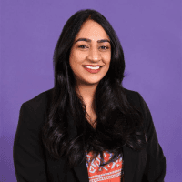A smiling person with long dark hair, wearing a black blazer, poses in front of a purple background.