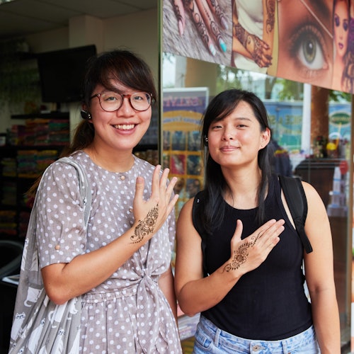 Two women proudly display intricate henna designs on their hands while standing outside a store with henna posters.