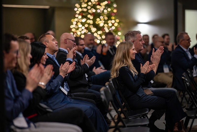 A group of people clapping and smiling at an event, with festive decorations in the background.
