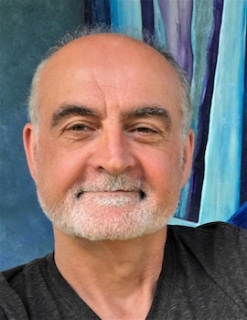 A smiling young man with crossed arms, wearing a plaid shirt and white t-shirt, poses against a dark background.