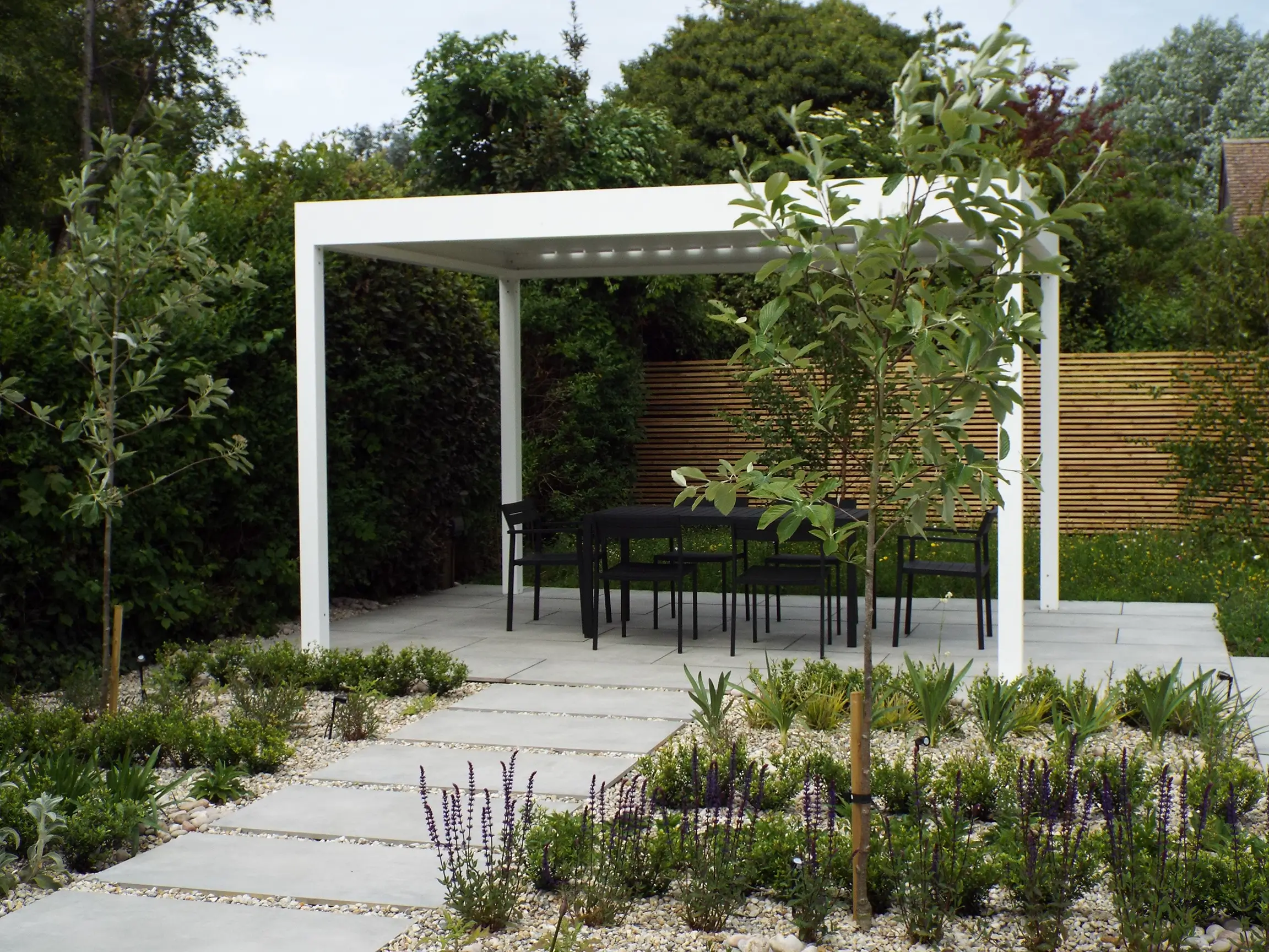 A modern outdoor pavilion with a seating area, surrounded by green plants and a stone pathway.
