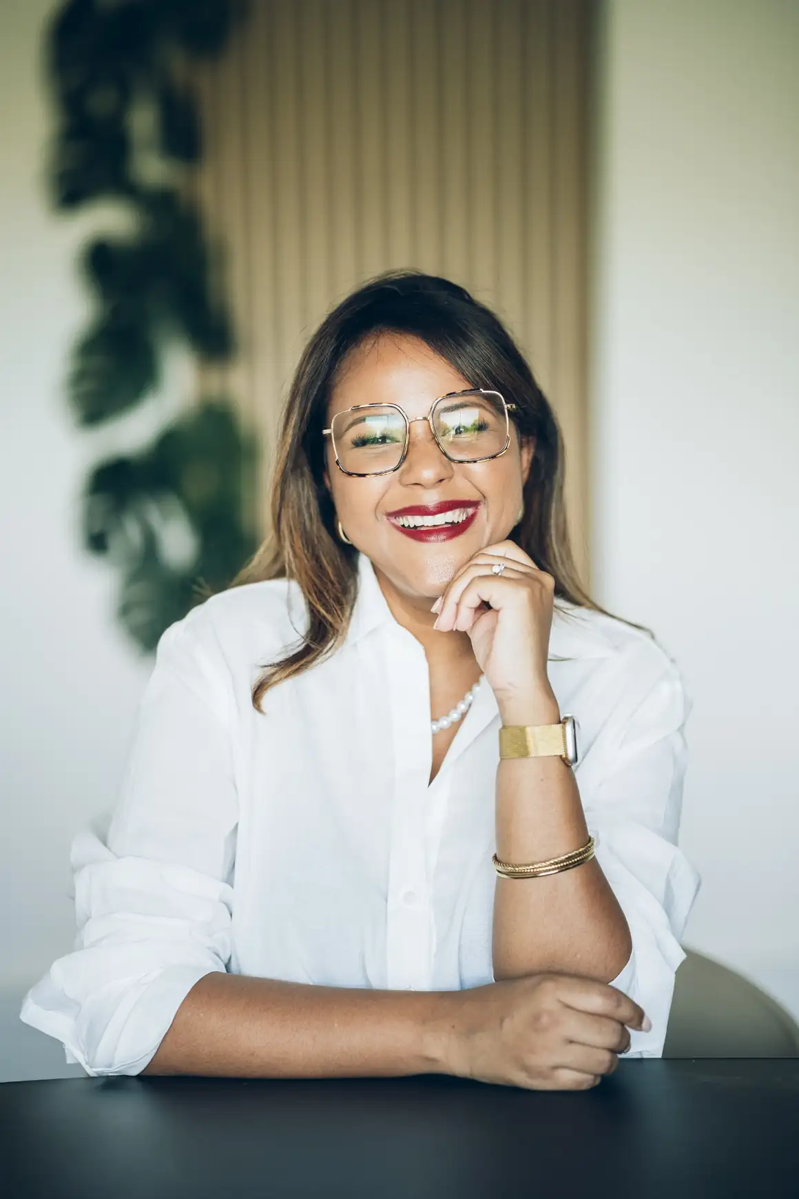 Portrait professionnel de Maître Julie Ramsamy souriante assise à un bureau, la tête appuyée sur la main, dans un environnement de travail lumineux.