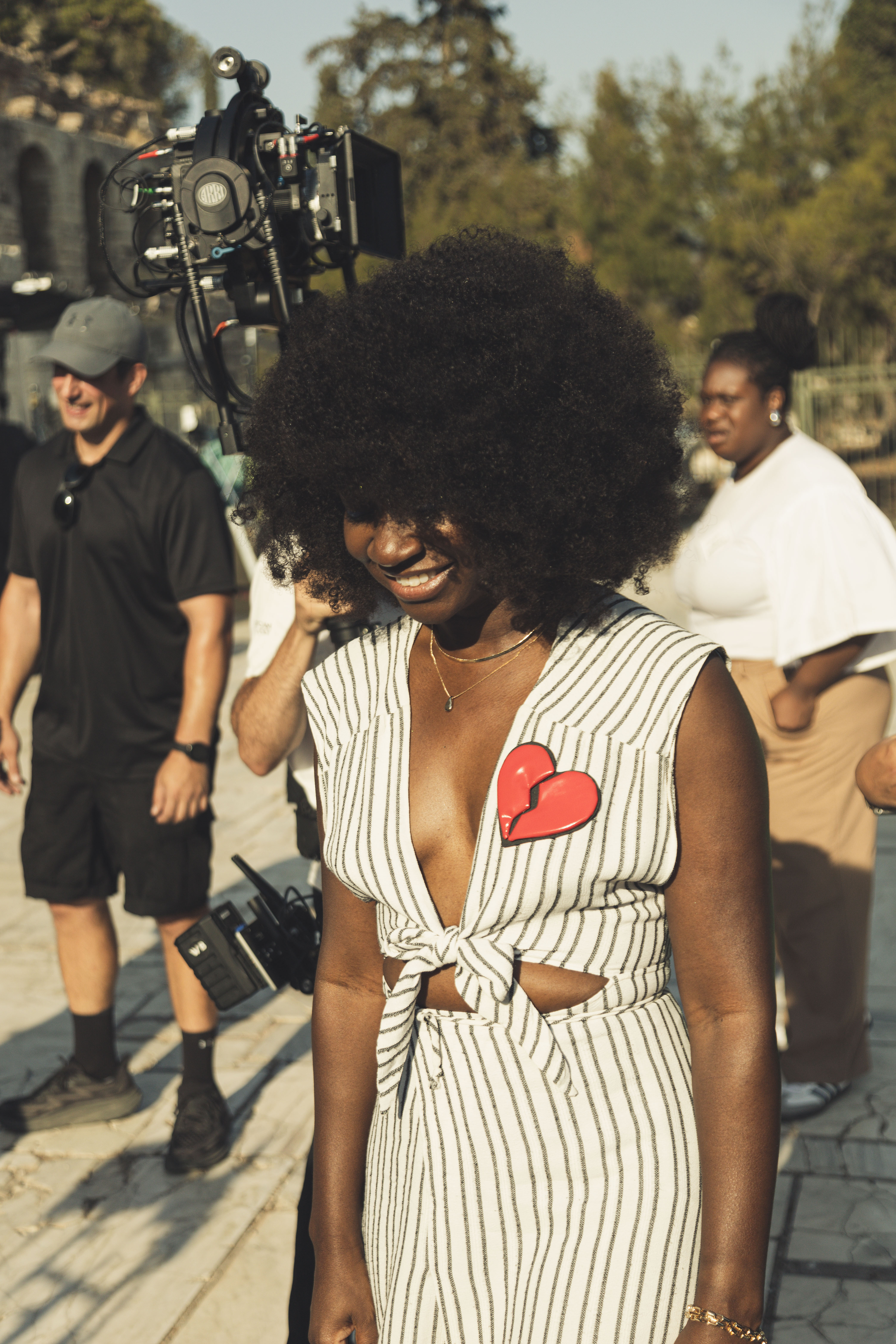 Natasha Ofili (a dark-skinned Black woman) with afro curly hair, a stripped jump-suit, and a large red heart pinned on her heart’s side that is slightly cracked down the middle but kept together.