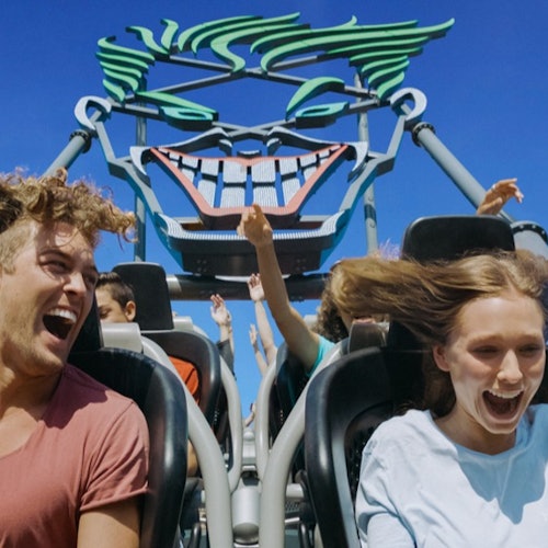 Twee mensen schreeuwen van vreugde in een achtbaan met een lachend gezichtontwerp op de achtergrond onder een heldere blauwe lucht.