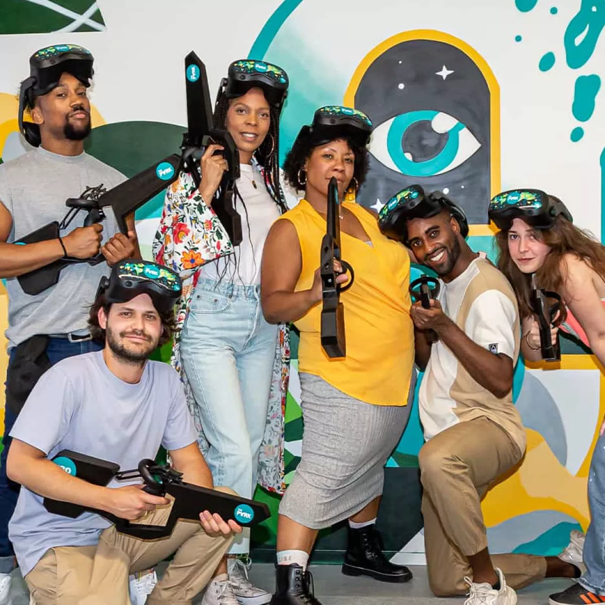 A diverse group of six friends and colleagues holding VR rifles and smiling, posing together in front of a colorful mural at The Park Cape Town.