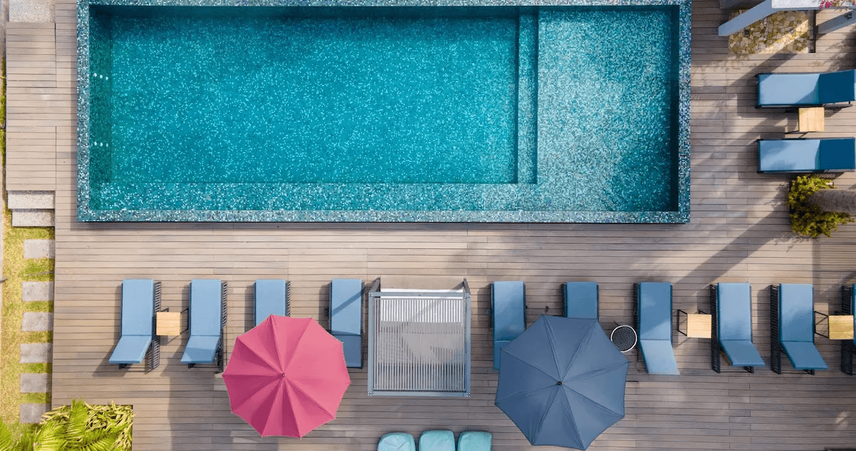 Aerial view of a modern rectangular swimming pool with surrounding timber deck, sun loungers, and colourful umbrellas.