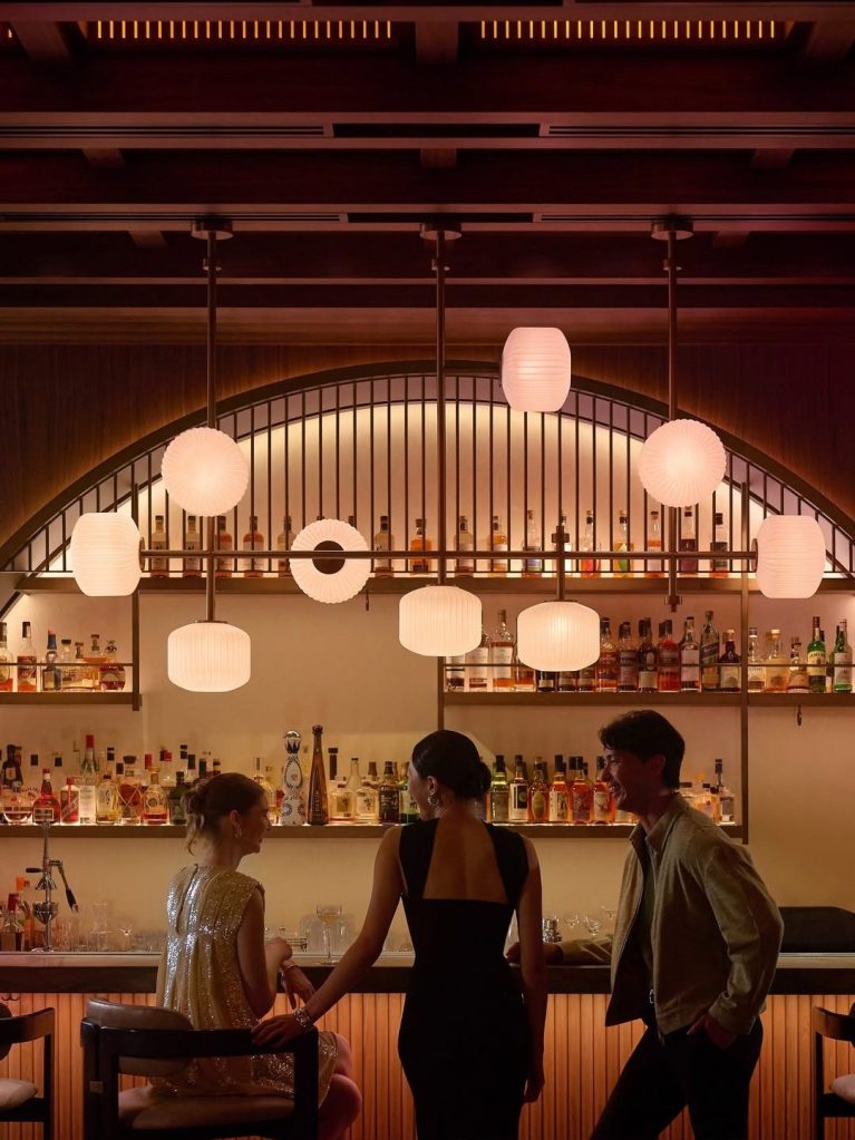 Interior of a stylish bar with a backlit shelf displaying various liquor bottles and elegant pendant lights. Two women in fashionable dresses converse, while a man stands nearby, smiling.