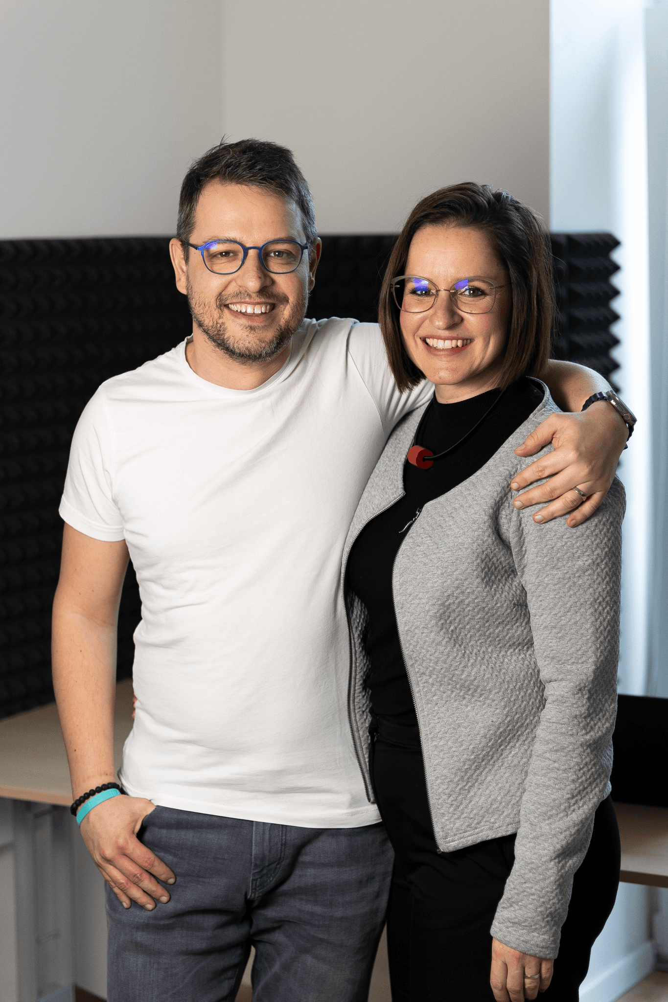 Dva usmívající se lidé pózují společně, jeden má na sobě bílou košili a druhý černý top a svetr.