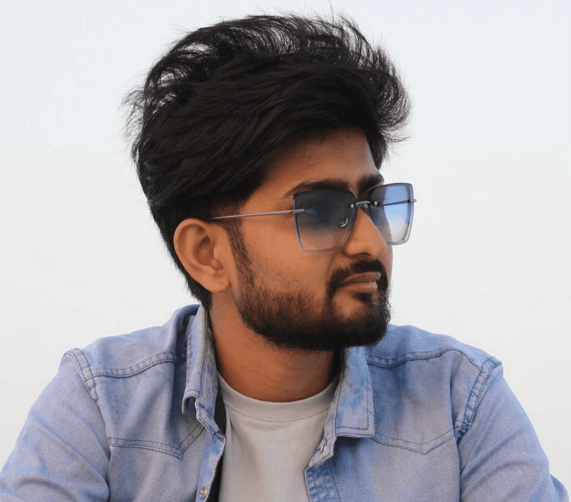 A man in a brown suit and cream silk scarf poses against a neutral background, exuding a composed and sophisticated demeanor.