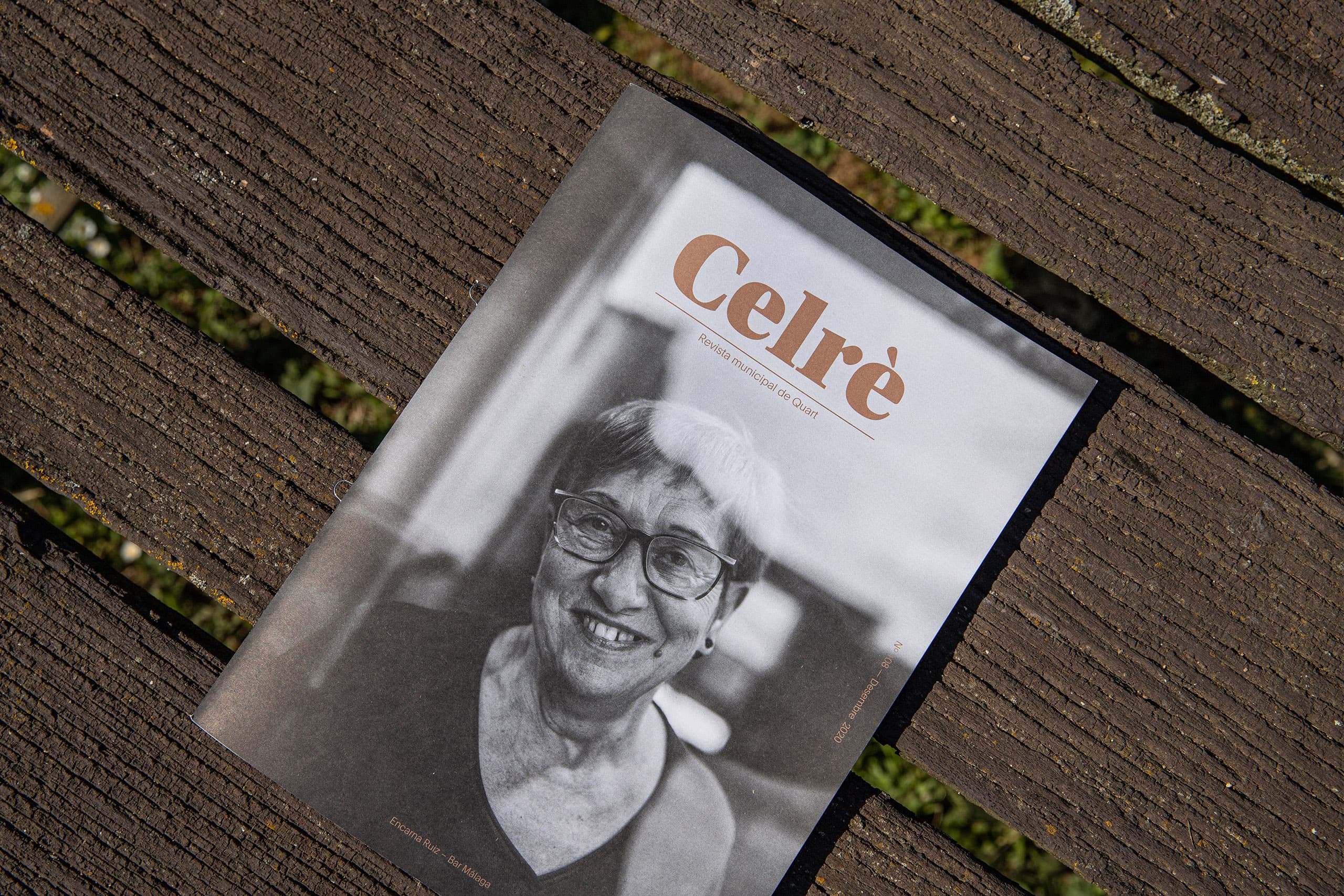 Overhead outdoor photograph showing a B&W cover of the "Celrè" magazine featuring a portrait of an smiling older woman with glasses