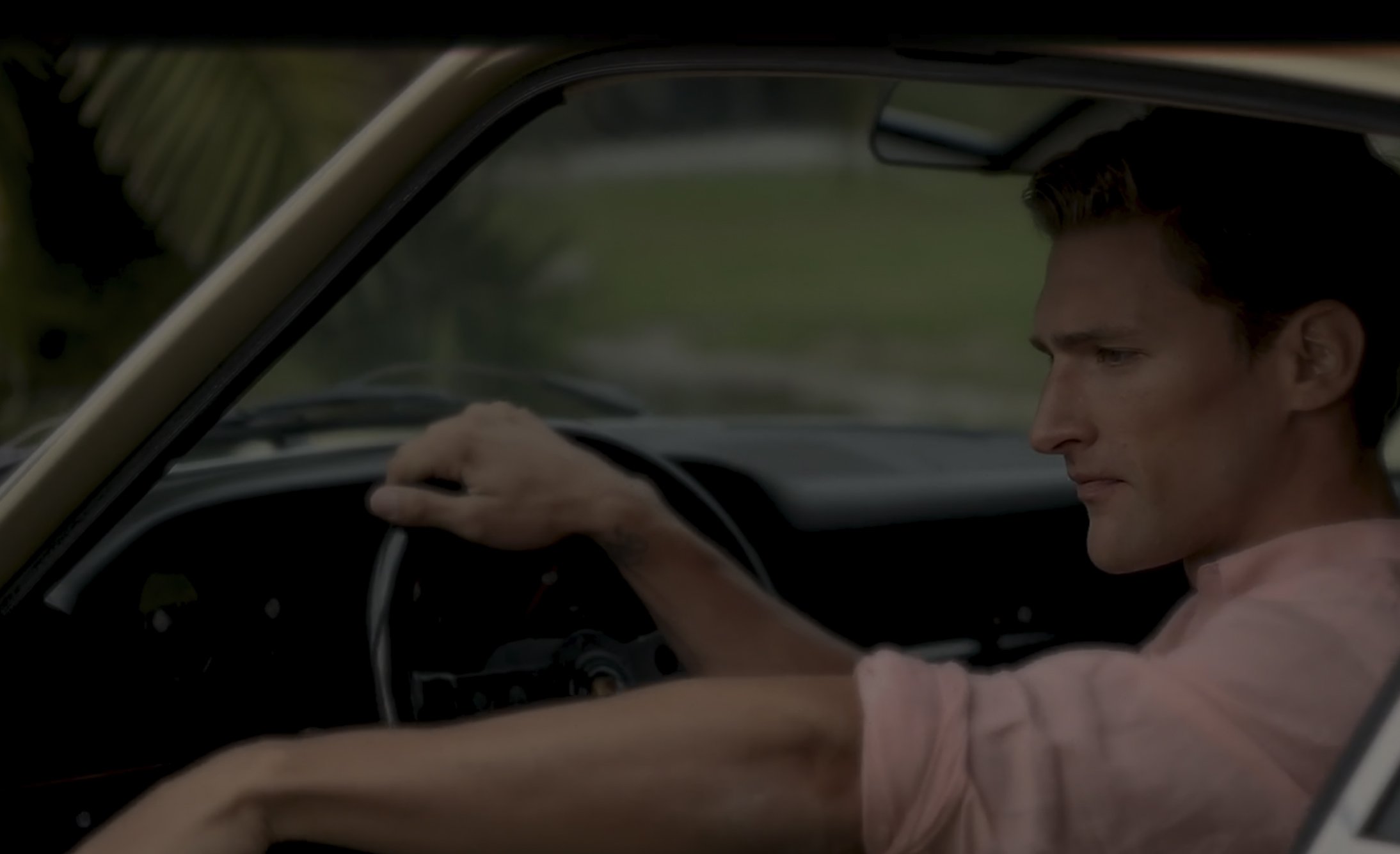 A man wearing a light pink shirt sits in the driver's seat of a car, gripping the steering wheel and looking forward with a serious expression.