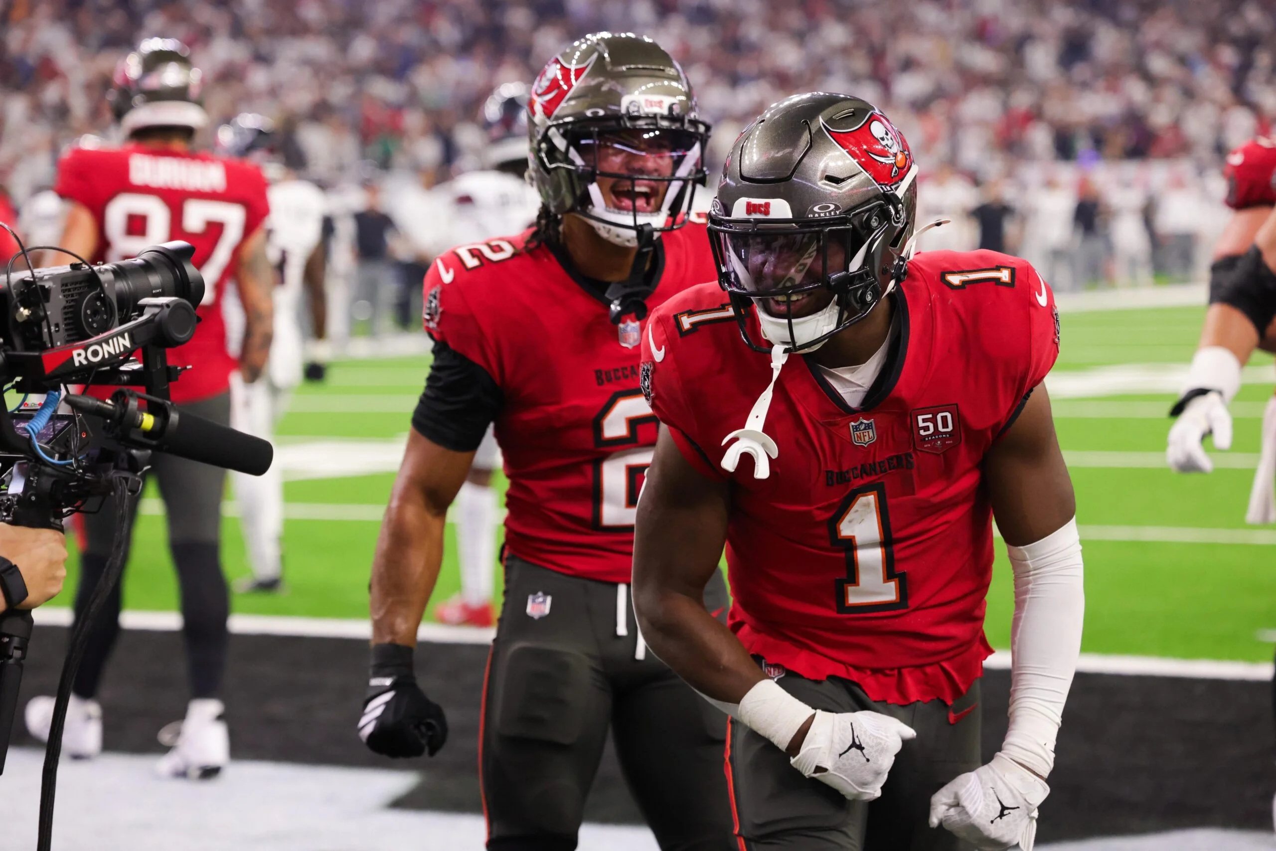 Tampa Bay Buccaneers players in red uniforms celebrate in the end zone during an NFL game, with one player flexing while a teammate shouts behind him on the field.