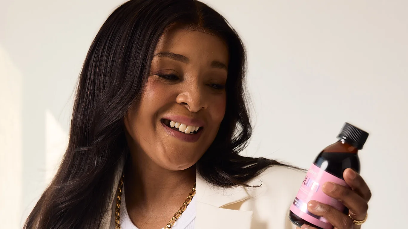 A woman in a cream suit smiles down at a bottle of Sēkret syrup in her hand