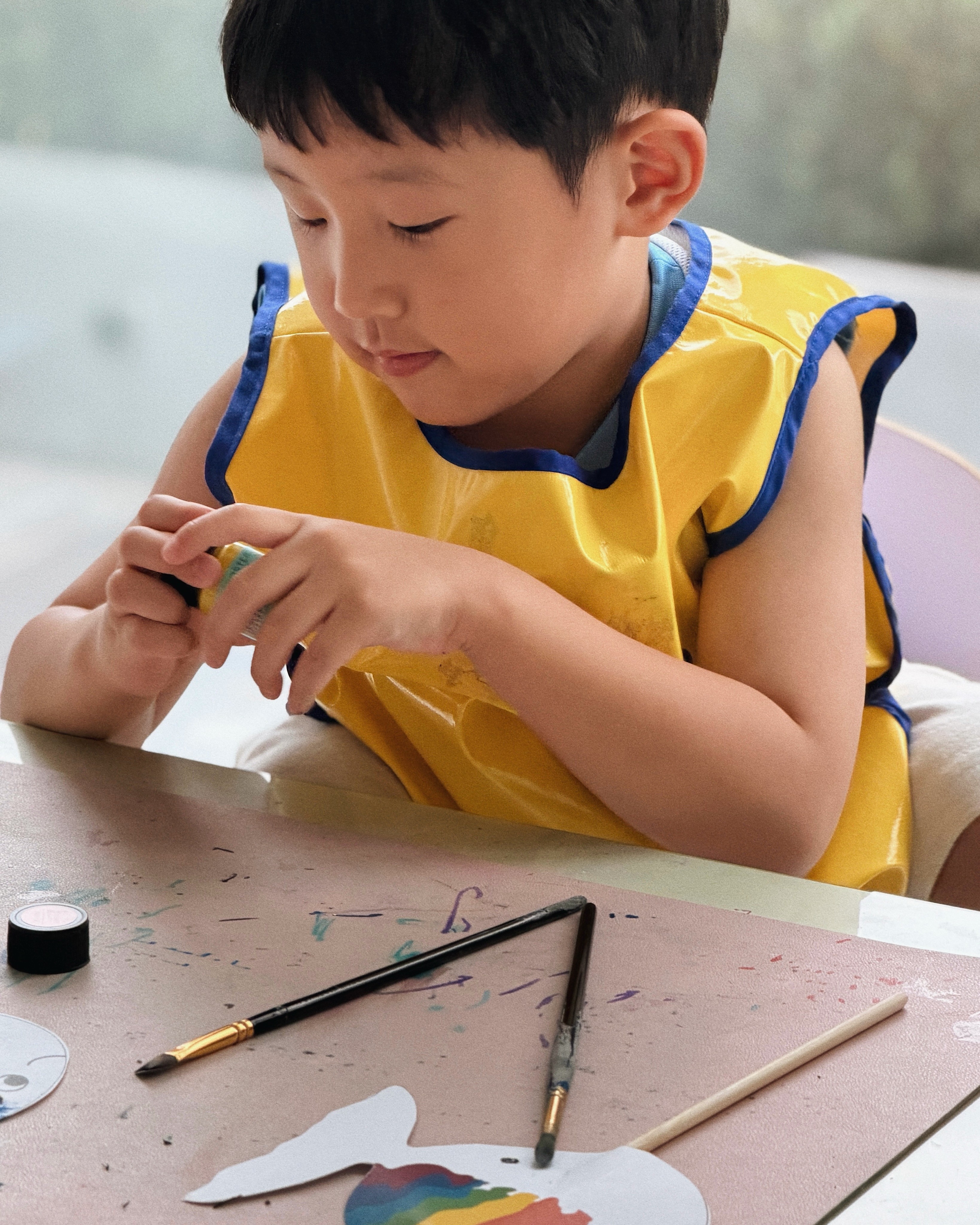 A young girl in a Noljak apron proudly holding a painted rainbow cardboard sword and a stick fish cutout from a storybook art class at Rookids BGC.