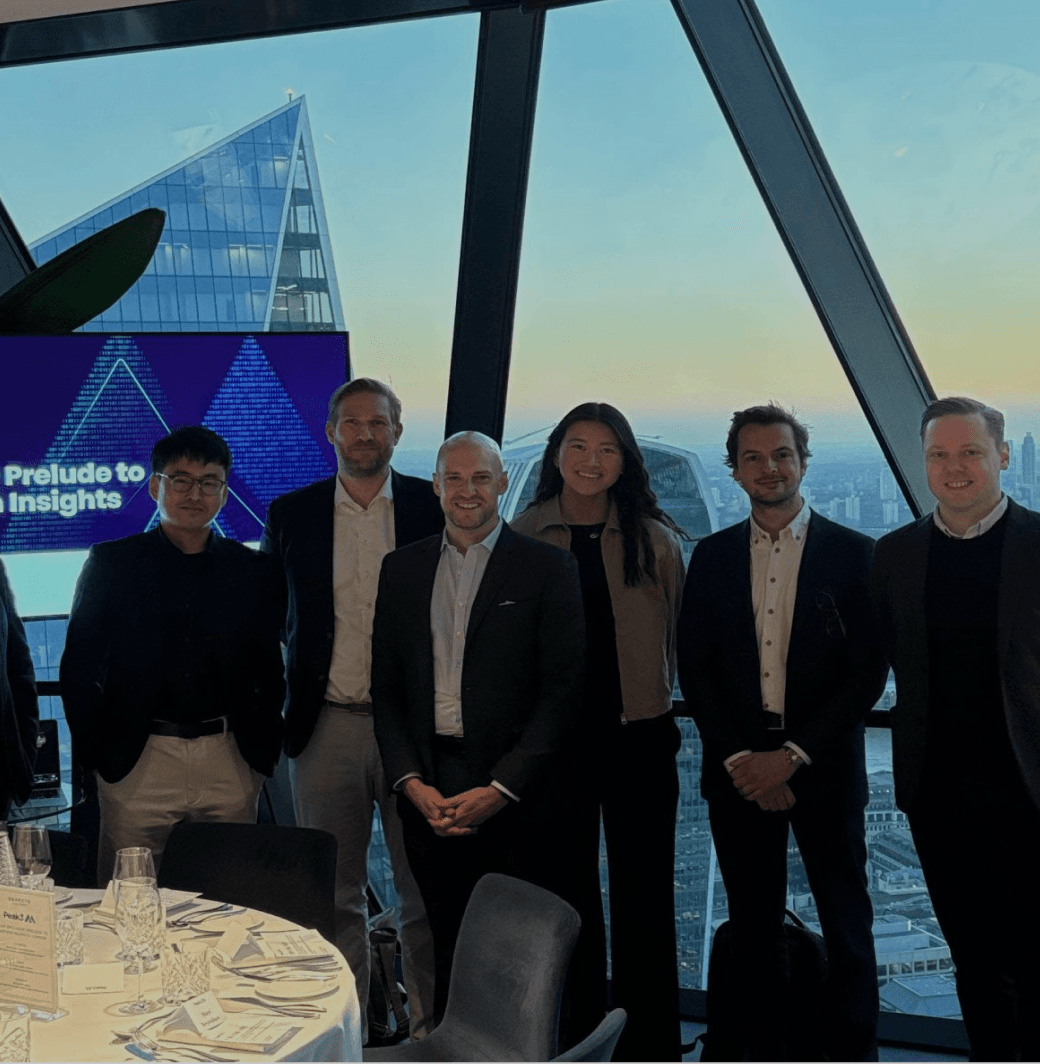 A group of six people standing together in a modern setting with a city view and a presentation screen behind them.